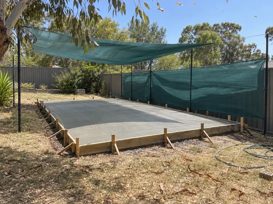 A freshly screeded concrete slab in timber formwork in a backyard on a hot, windy afternoon, with a shade-cloth windbreak tied along the side fence and more shade cloth stretched overhead.